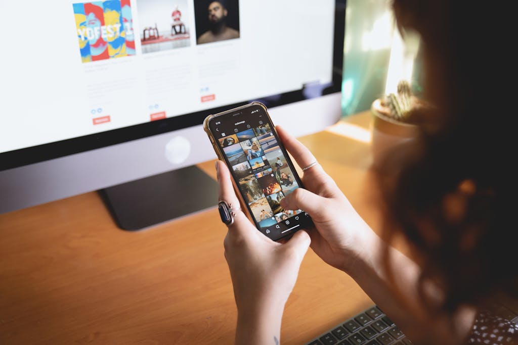 A person browsing images on a smartphone while sitting at a desk with a desktop monitor.